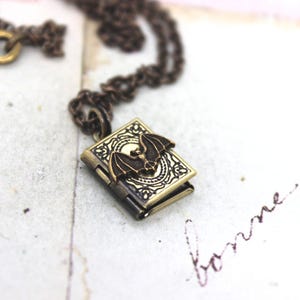 May include: A bronze-toned pendant necklace featuring a small, rectangular locket with a bat design. The locket is intricately detailed with an embossed pattern and opens to reveal a small compartment. The chain and clasp are also bronze-colored.