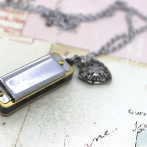 May include: A silver harmonica necklace with a heart-shaped pendant. The harmonica has the text "SWEET MUSIC" engraved on it. The necklace is made of a silver chain.