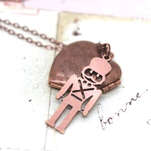 May include: A copper-colored necklace featuring a heart-shaped locket with a soldier charm. The locket is textured and opens to reveal a space for a small photo or keepsake. The soldier charm is a silhouette.