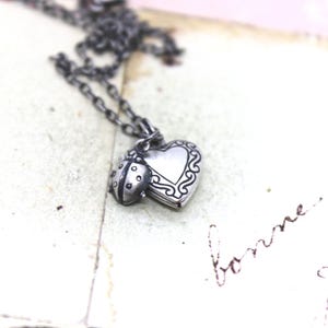 May include: A silver necklace with a heart-shaped locket and a ladybug charm. The locket has a delicate floral design.