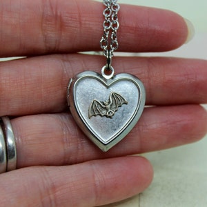 May include: A silver heart-shaped locket necklace with a silver chain. The locket has a small, gold-colored bat design on the front.