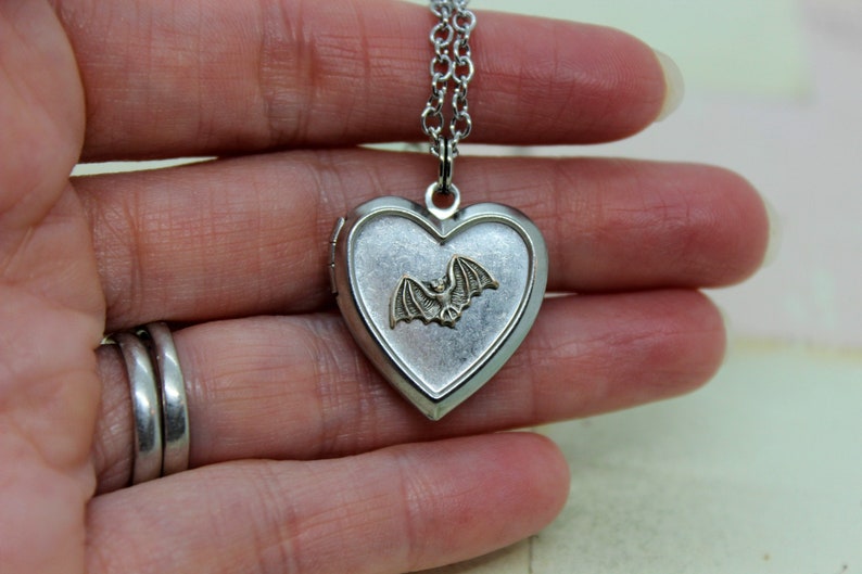 May include: A silver heart-shaped locket necklace with a silver chain. The locket has a small, gold-colored bat design on the front.