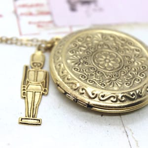May include: A gold-colored locket necklace with a detailed floral design and a small soldier charm. The round locket is approximately 1.5 inches in diameter and hangs from a delicate chain. The soldier charm is about 0.75 inches tall.
