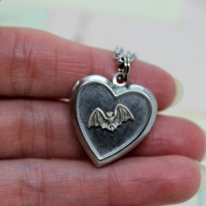 May include: Silver heart-shaped locket necklace with a silver bat charm inside. The locket has a dark gray background.
