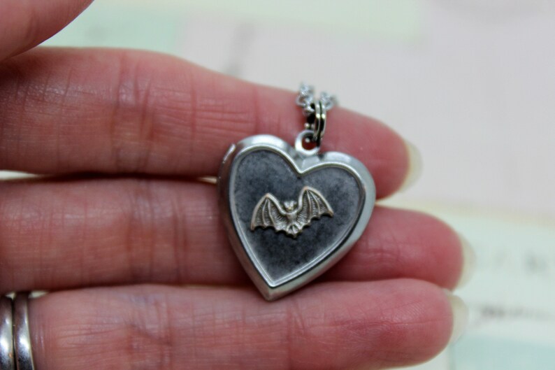 May include: Silver heart-shaped locket necklace with a silver bat charm inside. The locket has a dark gray background.