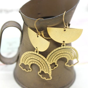 May include: Gold tone rainbow earrings with a half-circle design. The earrings are hanging from a brown ceramic pitcher.