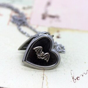 May include: A silver heart-shaped locket necklace with a black enamel finish and a silver bat design. The locket is open and the chain is visible.