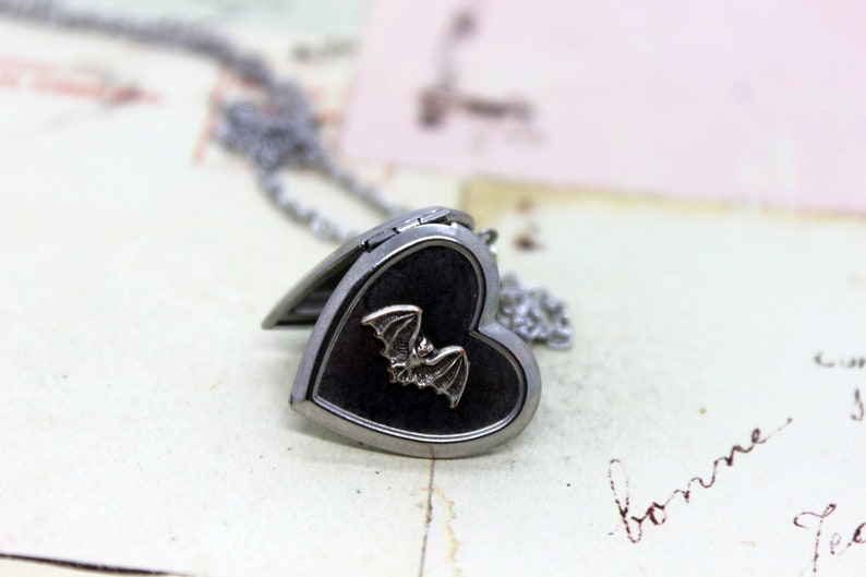 May include: A silver heart-shaped locket necklace with a black enamel finish and a silver bat design. The locket is open and the chain is visible.