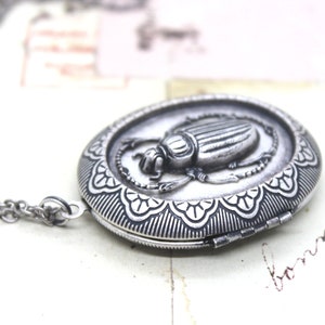 May include: Silver-toned oval locket pendant with a detailed scarab beetle design. The pendant is attached to a delicate chain. The locket features an ornate, patterned border. The jewelry is likely made of metal.
