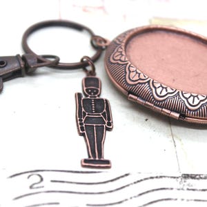 May include: A bronze-toned keychain featuring a decorative oval locket with an ornate border and a small soldier charm. The locket opens to reveal a space for a photo. The keychain includes a clasp for easy attachment.