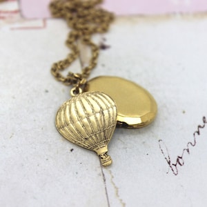 Puede incluir: Un collar de oro con un medallón y un dije de globo aerostático. El medallón está abierto y el dije de globo aerostático cuelga del medallón.