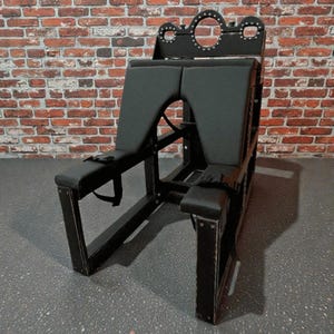 Puede incluir: Silla de madera negra con asiento y respaldo acolchados. La silla tiene un diseño único con recortes y correas. El marco es oscuro y la tapicería es negra. La silla está diseñada para un propósito específico.
