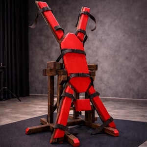 May include: A red wooden restraint device with black straps. The device is constructed with a wooden frame and features padded red sections for comfort. The straps are adjustable and secure. The device is designed for a specific purpose.