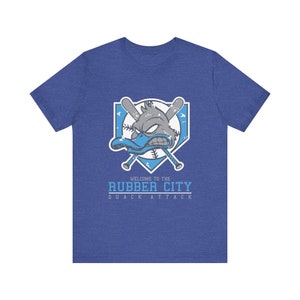 Può includere: Una maglietta blu con una grafica di un'anatra con una mazza da baseball e il testo "Welcome to the Rubber City Quack Attack".