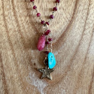 以下が含まれることがあります： 小さな赤いビーズのチェーンが付いたネックレス。ペンダントにはピンク色の石、ターコイズ色の石、金色の星のチャームが含まれています。ネックレスは木製の背景に表示されています。