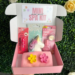 May include: A pink box filled with a mini spa kit. The kit includes a unicorn bath bomb, a lip balm, a nail polish, a nail polish remover, and a headband with two flower clips. The box has the text "Mini Spa Kit" on the top.