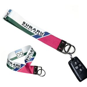 May include: A white, green, and pink Subaru Rally Team lanyard with a black key fob attached. The lanyard has the words "SUBARU RALLY TEAM" printed on it. A second, smaller lanyard is also visible.