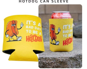 May include: Yellow hotdog can sleeve with a cartoon hotdog illustration. The text reads "IT'S A BAD DAY TO BE A HOTDOG". The can sleeve is designed to keep beverages cold and features a black trim.