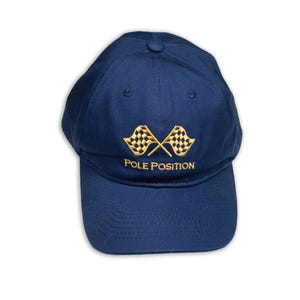 Puede incluir: Gorra de béisbol azul marino con visera curva. La gorra presenta un diseño bordado de dos banderas a cuadros doradas cruzadas, con las palabras "POLE POSITION" bordadas debajo.