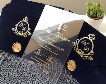 Könnte beinhalten: Eine transparente Acryl-Hochzeitseinladung mit goldenen Lettern und einem goldenen Monogramm. Die Einladung liegt auf einem dunkelblauen Samt-Umschlag mit einem goldenen Siegel.