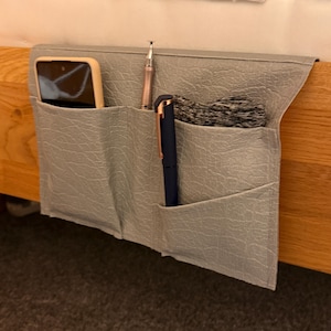 May include: A grey fabric organiser with three pockets, hanging on the side of a bed. The organiser has a mobile phone, a pen, and a pencil inside.