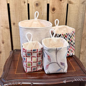May include: A set of four fabric storage baskets with hanging loops. One large basket is solid beige, one medium basket features a colorful chevron pattern, and two smaller baskets have geometric square and leaf-like floral patterns respectively.