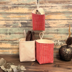 May include: Three hanging fabric storage baskets. Two are red with vertical white stripes, and one is off-white. The baskets have a loop at the top for hanging. One basket contains brown items.