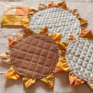 May include: A collection of quilted fabric coasters and mats shaped like sunflowers. The set includes a square coaster, two round mats with a light blue and white gingham pattern, and one round mat with a textured brown fabric centre.