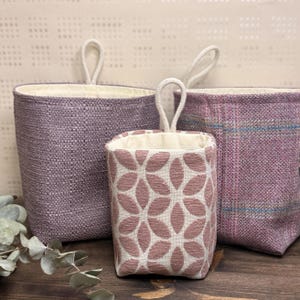May include: Three fabric storage baskets in shades of purple. The baskets have a woven texture and cream-coloured interiors. The smallest basket features a floral pattern in pink and cream. Each basket has a loop handle made of a cream-coloured rope.