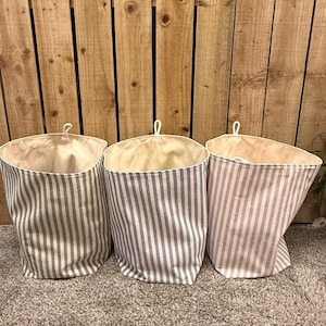 May include: Three fabric storage baskets with vertical stripes in grey, blue, and lavender. Each basket has a cream-coloured interior and a loop for hanging. Ideal for organizing items, these baskets are a practical storage solution.