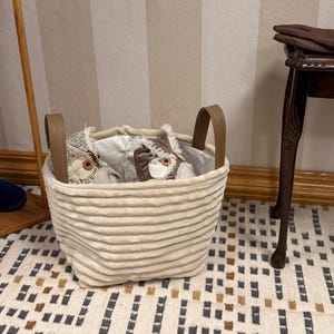 May include: A beige, corduroy-textured storage basket with brown leather handles. The basket contains two stuffed owl toys with large, round eyes. The basket sits on a patterned rug, with a wooden table and gloves in the background.