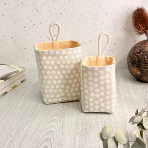 May include: Two rectangular fabric storage baskets with a beige and white polka dot pattern. Each basket features a light peach interior lining and a single fabric loop handle attached to the top center for hanging or carrying purposes.