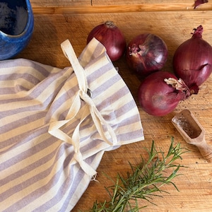 May include: A striped lavender and white fabric bag with a drawstring closure, next to several red onions, rosemary sprigs, and a small wooden scoop of salt on a wooden cutting board. A blue ceramic bowl is in the background.