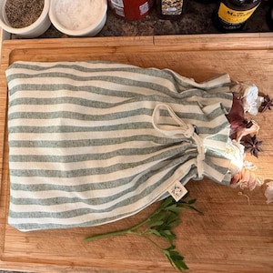 May include: A green and white striped drawstring bag on a wooden chopping board. The bag contains garlic and star anise. A sprig of parsley is in the foreground. The bag has a small tag with a sun design.