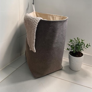 May include: A grey fabric laundry basket with a beige lining. A cream-coloured knitted item hangs over the side. A small potted plant with green leaves is nearby. The basket is designed to fit in a corner.