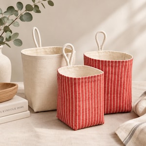 May include: Three fabric storage baskets with hanging loops. One basket is solid off-white, while two feature a red and white vertical striped pattern. Each basket has a structured, upright design with a light-colored interior lining and a flat base.