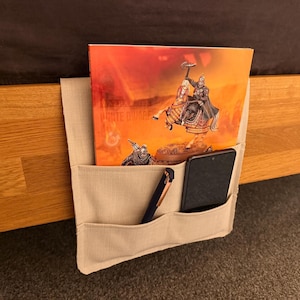 May include: Beige canvas bed organiser with three pockets. The organiser is hanging on the side of a wooden bed frame. A phone, pen, and a book with a fantasy illustration are in the pockets.