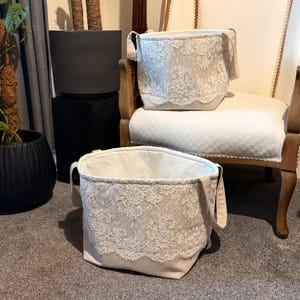 May include: Two beige fabric storage baskets with white lace trim. The baskets have handles and are sitting on a chair and the floor.