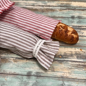 May include: Two striped bread bags, one red and white, the other brown and white, with a baguette in one. The brown bag has a white lace trim. The bags are on a weathered, turquoise and brown wooden surface.
