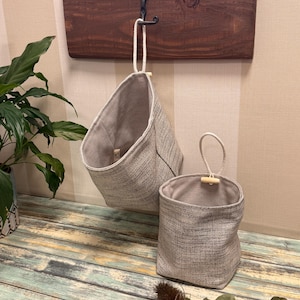 May include: Two gray fabric hanging baskets with a textured weave. The larger basket hangs from a wooden board with a metal hook. The smaller basket has a loop handle with a wooden toggle. The baskets are lined with a lighter gray fabric.