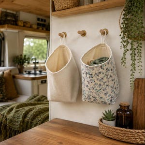 May include: Two hanging fabric storage baskets suspended from wooden wall hooks. One basket is solid off-white, while the other features a blue and green floral pattern. Both baskets have a wide opening and a loop for hanging on the wall.