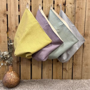 May include: Four triangular fabric storage pockets hanging in a row, featuring a textured weave in lime green, lavender, sage green, and light grey. Each pocket has a cream-coloured interior lining and a small fabric loop for hanging on a hook.