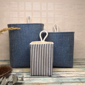 May include: Three fabric storage containers. Two are a solid blue with white rope handles. The third is a smaller, rectangular container with vertical navy and white stripes and a white rope handle. The containers are on a wooden surface.