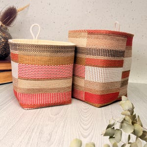 May include: Two rectangular storage baskets featuring a woven pattern in shades of red, beige, brown, and cream. Each basket has a light-coloured fabric interior and a small white rope loop attached to the top edge for hanging purposes.