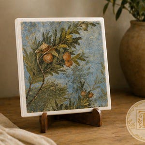 May include: A square ceramic tile featuring a detailed botanical illustration of pomegranate branches with orange fruit and green leaves against a textured blue background. The tile is displayed on a dark brown wooden stand with a white border.