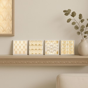 May include: Four square coasters with Art Deco-style gold patterns on a cream background. The coasters are arranged on a decorative shelf, with a framed artwork and a vase with greenery in the background. The patterns vary slightly across each coaster.