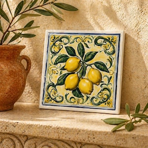 May include: A square ceramic tile featuring a hand-painted illustration of three yellow lemons with green leaves on a pale yellow background. The design is framed by intricate green and yellow scrollwork patterns and a thin blue border line.