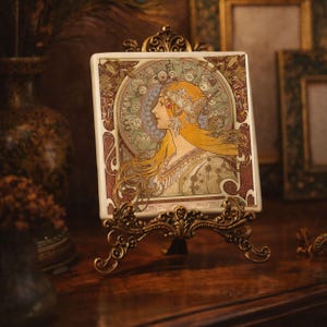May include: A decorative ceramic tile with an Art Nouveau illustration of a woman with long blonde hair, displayed on a brass easel. The artwork has a circular design with astrological symbols and is framed by ornate details. The colour palette includes gold, cream, and burgundy.