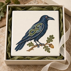 May include: A square white ceramic tile featuring a dark blue raven with a green and blue Celtic knot pattern on its wing. The bird is perched on a brown oak branch with green leaves and a single brown acorn, presented in a gift box.