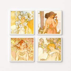 May include: A set of four square coasters featuring Art Nouveau style illustrations of women surrounded by flowers. The designs include lilies, a red flower, roses, and irises in soft pastel tones of yellow, pink, peach, and light blue.